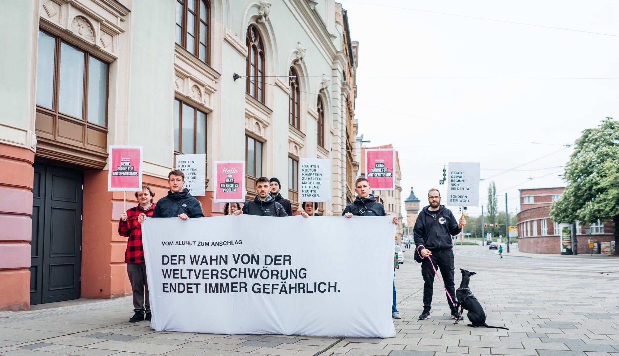 Personen mit Schildern in den Händen stehen hinter einem Transparent mit der Aufschrift "Vom Aluhut zum Anschlag: Der Wahn von der Weltverschwörung endet immer gefährlich. Die protestierende Personengruppe steht vor dem Steintor-Varieté in Halle (Saale).